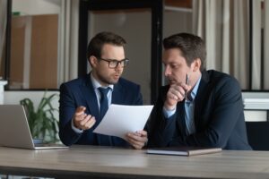 A lawyer and their client in suits sitting at a table next to each other, with the lawyer holding a piece of paper and discussing Massachusetts labor laws for salaried employees.