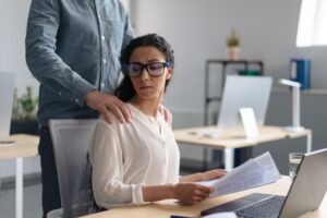 A young female at a desk being touched on the shoulders by a male coworker wonders what is considered sexual harassment at work.