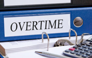 The word "overtime" appears on a piece of paper attached to the spine of a blue binder. If Costco has not paid you for the overtime you worked, consult an employment attorney who can file a complaint and take legal action to recover your wages.